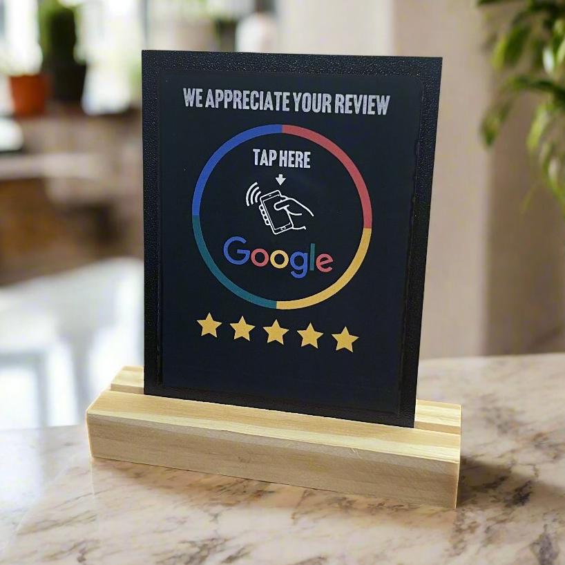 A black NFC Tap Card with a Google logo and a prompt to 'Tap here to leave a Google review' placed on a wooden stand, with a 5-star rating icon below it.